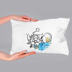 May include: A white pillow with a black line drawing of a teapot with blue and yellow flowers.