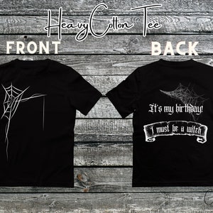 Pode incluir: T-shirt preta de algodão pesado com um desenho de teia de aranha branca na frente e atrás. A parte de trás da camisa tem o texto "It's my birthday! I must be a witch" em uma fonte gótica. A camisa é exibida em um fundo de madeira.