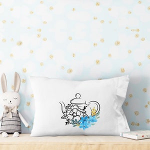 May include: White pillow with a black line drawing of a teapot with blue flowers. The pillow is on a light blue background with gold glitter.