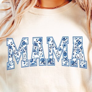 May include: A white t-shirt with a blue floral print that says "MAMA" in a varsity style font.