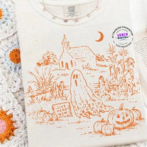 May include: A cream-colored t-shirt featuring an orange line-art graphic of a church, a ghost, pumpkins, and corn stalks. The text "LIVE HERE NOW" is printed on a small sign. A circular logo with the text "AI TRAINING PROHIBITED ARWEN DIGITAL" is visible.