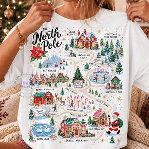 May include: A white t-shirt featuring a colorful map of the North Pole. The graphic includes illustrations of Santa's workshop, reindeer stables, an elf village, and candy cane lanes, with text labels for each location and a central North Pole sign.