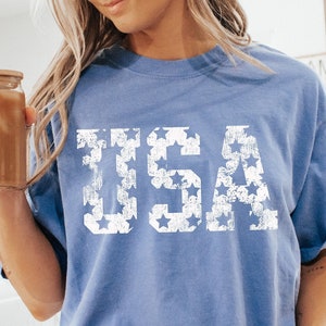 May include: A woman wearing a light blue t-shirt with a white "USA" graphic made of stars. The shirt is slightly oversized and has a relaxed fit. The woman is sitting on a white surface with her legs crossed. She is wearing white socks with black stripes.