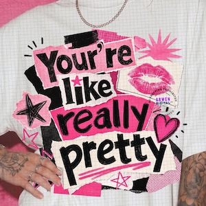 May include: A white t-shirt featuring a grid pattern and a graphic design with the text "You're like really pretty" in black and pink. The design includes a pink lip print, black stars, and a pink heart, layered over a collage of paper-like shapes.
