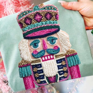 May include: A light green t-shirt with an embroidered nutcracker design. The nutcracker has a detailed hat, uniform, and mustache, with pink, teal, gold, and blue accents. The shirt is held up in front of a pink Christmas tree, with festive decorations.