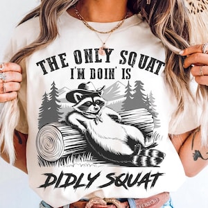 May include: Cream-colored t-shirt with a graphic of a raccoon wearing a cowboy hat, lounging on a log. The text reads "THE ONLY SQUAT I'M DOIN' IS DIDLY SQUAT" in black. The design includes a mountain and forest scene.