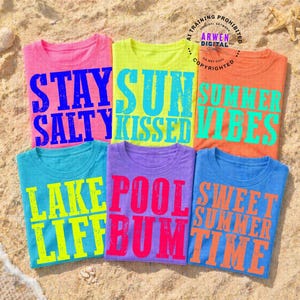 May include: Six colorful t-shirts with summer-themed phrases printed on them. The shirts are arranged on a sandy surface. Phrases include "Stay Salty", "Sun Kissed", "Summer Vibes", "Lake Life", "Pool Bum", and "Sweet Summer Time".