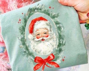 May include: A light blue sweatshirt featuring a vintage Santa Claus portrait within a green wreath and a red bow. The Santa has a white beard and a red hat. The sweatshirt is held up in front of a pink Christmas tree.