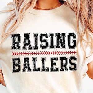 May include: A white t-shirt with the text "Raising Ballers" in black with a red baseball stitch design.