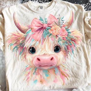May include: Cream-colored t-shirt featuring a colorful illustration of a Highland cow. The cow has large blue eyes, a pink nose, and is adorned with a pink bow, flowers, and colorful accents. The shirt is made of a soft material.