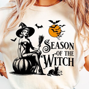 May include: Cream-colored t-shirt with a black and white graphic featuring a witch sitting on a pumpkin, bats, and the text "Season of the Witch." A black cat and skull are also in the design. The shirt has short sleeves.