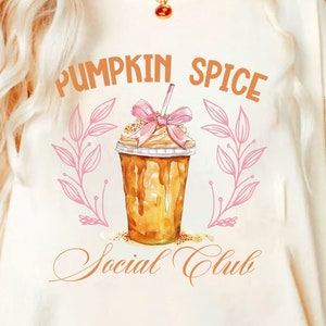 May include: Cream-colored t-shirt featuring the text "PUMPKIN SPICE Social Club" in orange script. A watercolor illustration of a pumpkin spice latte with a pink bow and leaves is centered on the shirt.