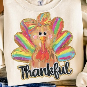 May include: Cream-colored sweatshirt featuring a colorful turkey design with a rainbow-colored tail and the word "Thankful" in black script. The sweatshirt is paired with blue denim shorts, a tan hat, and sandals, creating a casual, festive look.