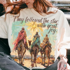 May include: Cream-colored t-shirt with a graphic of three figures on camels, with the text "They followed the star with great joy." The image has a watercolor style with warm tones. The person in the image is wearing a brown cap and sunglasses.