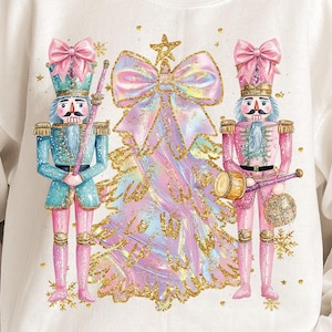 May include: White sweatshirt with a Christmas tree and two nutcracker soldiers. The tree is decorated with a large pink bow and gold glitter. The nutcrackers are wearing pink and blue uniforms and holding musical instruments.