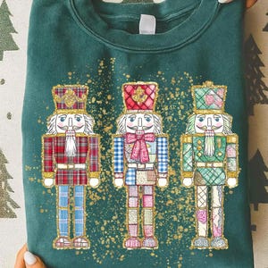May include: A dark green sweatshirt featuring three whimsical nutcracker figures. Each nutcracker has a unique design with plaid and patchwork patterns in red, blue, and green. Gold accents and a gold splatter design surround the figures, creating a festive holiday theme.