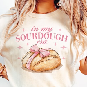 May include: A cream-colored t-shirt with the text "in my sourdough era" in pink lettering. A cartoon illustration of a loaf of sourdough bread with a pink ribbon is below the text.