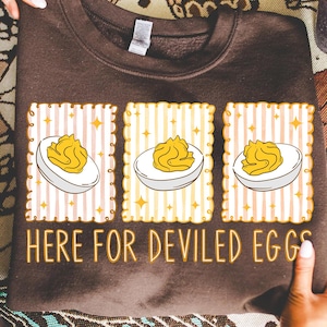 May include: A brown sweatshirt with the text "HERE FOR DEVILED EGGS." The graphic features three deviled eggs in decorative frames with striped and star backgrounds. The sweatshirt is held up by hands.