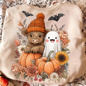 Può includere: Felpa beige con un design a tema Halloween. Presenta una mucca delle Highlands con un cappello arancione, un fantasma amichevole, zucche, fiori e pipistrelli. Il design è nei caldi colori autunnali.