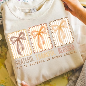 May include: A beige sweatshirt featuring three rectangular designs with bows in shades of brown and orange. The text "Grateful Thankful Blessed" is printed below the designs, with the phrase "God is faithful in every season."