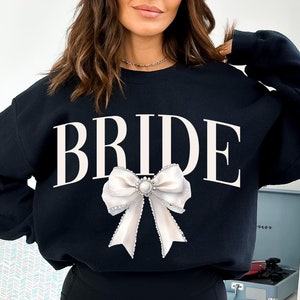 May include: Black sweatshirt with the word "BRIDE" printed in white and a white satin bow with a pearl brooch.
