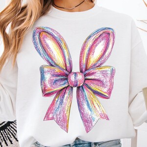 May include: A white sweatshirt with a large, colorful, glittery bow design on the front. The bow is pink, purple, yellow, and blue.