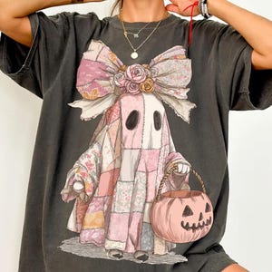 May include: Dark gray oversized t-shirt featuring a whimsical ghost design. The ghost is depicted in a patchwork quilt costume, holding a pumpkin basket. A large pink bow with floral accents adorns the ghost's head, perfect for Halloween.