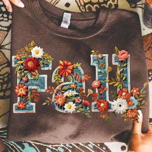 May include: A brown sweatshirt with the word "FALL" embroidered in a floral design. The letters are outlined in a teal color and filled with colorful flowers in shades of red, orange, and white.