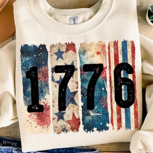 Puede incluir: Una sudadera de color crema con un diseño de bandera estadounidense vintage que presenta el número 1776 en negro. El diseño incluye estrellas y fuegos artificiales rojos, blancos y azules.