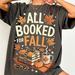 May include: Dark gray t-shirt with the text "ALL BOOKED FOR FALL" and fall-themed graphics. The design includes books, a pumpkin, a coffee mug, and autumn leaves. The shirt is a relaxed fit.