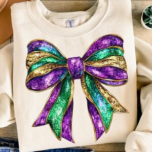 May include: A cream-colored sweatshirt with a large purple, green, and gold glitter bow on the front. The bow has a sparkly, iridescent finish.
