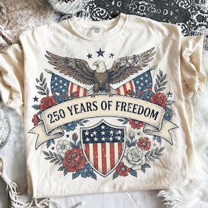 Könnte beinhalten: Cremefarbenes T-Shirt mit patriotischem Design. Die Grafik zeigt einen Adler mit ausgebreiteten Flügeln, amerikanische Flaggen, einen Schild und den Text "250 YEARS OF FREEDOM" auf einem Banner. Blumenelemente umgeben das Design.