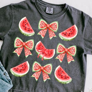 May include: A dark grey t-shirt with a watermelon and bow print. The watermelon slices are red and green with black seeds. The bows are red with white polka dots.