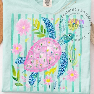 May include: A light blue t-shirt with a watercolour design of a sea turtle. The turtle has a pink shell and blue flippers, surrounded by pink flowers and green foliage, set against a striped background.