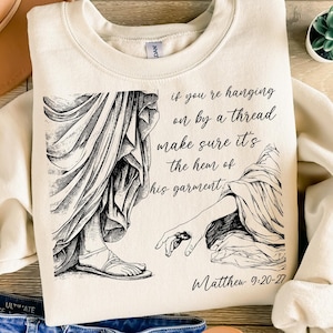 May include: A cream-colored sweatshirt with a black and white illustration of a hand reaching out from a robe. The text "if you're hanging on by a thread make sure it's the hem of his garment" is printed on the sweatshirt, along with the scripture reference "Matthew 9:20-22".