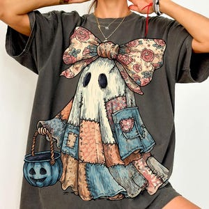 Può includere: T-shirt grigio scuro con un fantasioso disegno di fantasma. Il fantasma è adornato con un corpo patchwork, un fiocco floreale e un secchio di zucca blu. Il design ha un'estetica vintage e artigianale, perfetto per Halloween.