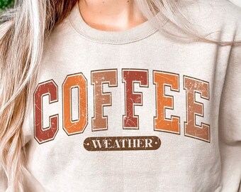 May include: A beige sweatshirt with the words "COFFEE WEATHER" in a retro, collegiate-style font. The text is in shades of brown and orange. The sweatshirt has a crew neck and long sleeves.