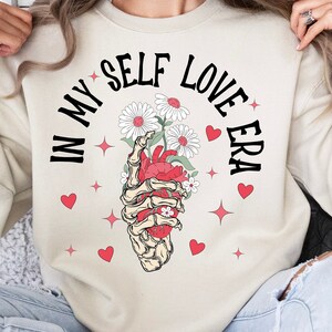 May include: Cream-colored sweatshirt with the text "IN MY SELF LOVE ERA" in black. The graphic features a skeletal hand holding a heart with flowers, surrounded by red hearts and stars. A cozy and stylish top.