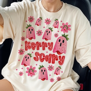 Puede incluir: Camiseta color crema con un diseño lúdico de fantasmas y flores de fresa rosas. El texto "Berry Scary" está escrito en rojo. El diseño incluye estrellas y un tema caprichoso y espeluznante.