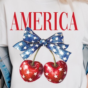 May include: White t-shirt with the word "AMERICA" printed in red letters. A red and blue bow with white stars is centered below the word. Two red cherries with white stars are below the bow.