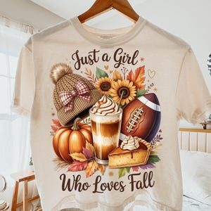 K&ouml;nnte beinhalten: Cremefarbenes T-Shirt mit einem Herbst-Themen-Motiv. Das Design umfasst eine Strickm&uuml;tze, einen K&uuml;rbis, Sonnenblumen, einen Football, ein Kaffeegetr&auml;nk und ein St&uuml;ck Kuchen. Der Text lautet "Just a Girl Who Loves Fall."