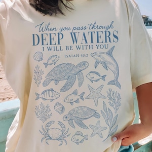 Puede incluir: Camiseta color crema con un diseño azul de criaturas marinas y el texto "When you pass through DEEP WATERS I WILL BE WITH YOU ISAIAH 43:2". El diseño incluye tortugas, peces, estrellas de mar y un delfín.