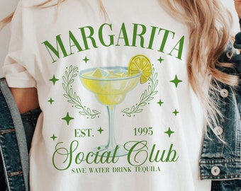 May include: White t-shirt with a green graphic design that says "Margarita Social Club Est. 1995 Save Water Drink Tequila" with a drawing of a margarita glass with a lime wedge.