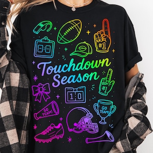 Puede incluir: Camiseta negra con un gráfico colorido de iconos relacionados con el fútbol, incluyendo un balón, un casco y un trofeo. El texto "Touchdown Season" está escrito en una fuente de guion. El diseño presenta un efecto de gradiente arcoíris.