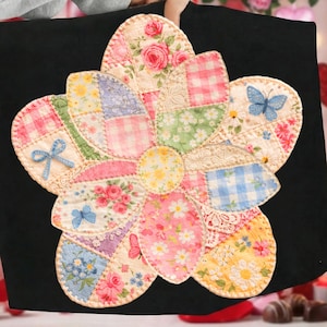 May include: A black tote bag featuring a large, appliqued flower design. The flower is composed of various fabric patches in pastel colors, including pink, blue, and yellow, with floral and gingham patterns. A blue bow and butterfly accents add detail.