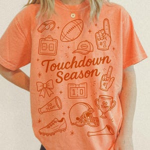 May include: Orange t-shirt with the text "Touchdown Season" and football-themed graphics. The design includes a football, helmet, and a trophy. The shirt is a comfortable fit.