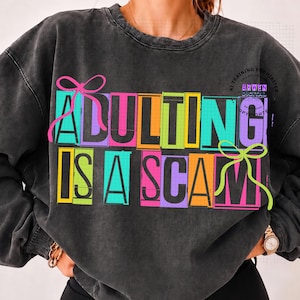 May include: A charcoal gray crewneck sweatshirt featuring the text ADULTING IS A SCAM in bold, blocky letters. Each letter is set against a colorful rectangular background in shades of pink, yellow, orange, teal, and purple, with pink and lime green bows.