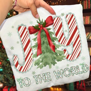 May include: White Christmas-themed pillow with the words "JOY TO THE WORLD" in red and green. The "JOY" letters have a candy cane stripe design with a green lace border. A watercolor Christmas tree and red bow are in the center.