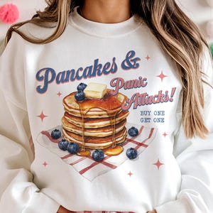 以下が含まれることがあります： 白いスウェットシャツに、パンケーキの積み重ねと「Pancakes & Panic Attacks! Buy one get one」というテキストのグラフィックがプリントされています。モデルはピンクと白のチェック柄のショートパンツを履いています。