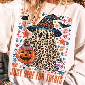 May include: A white crewneck sweatshirt featuring a graphic of a ghost with a leopard print pattern wearing a witch hat. The ghost holds an orange jack-o'-lantern bucket filled with candy. The text "JUST HERE FOR TREATS" is printed below.